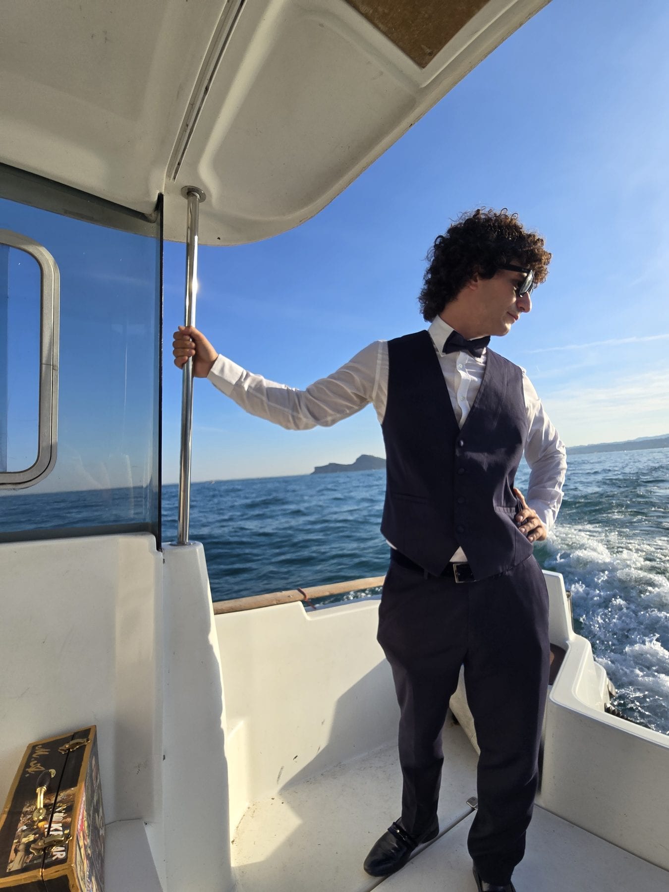 Matteo Angrisano in gilet e papillon a bordo della barca che raggiunge l'isola, con la valigia da lavoro accanto