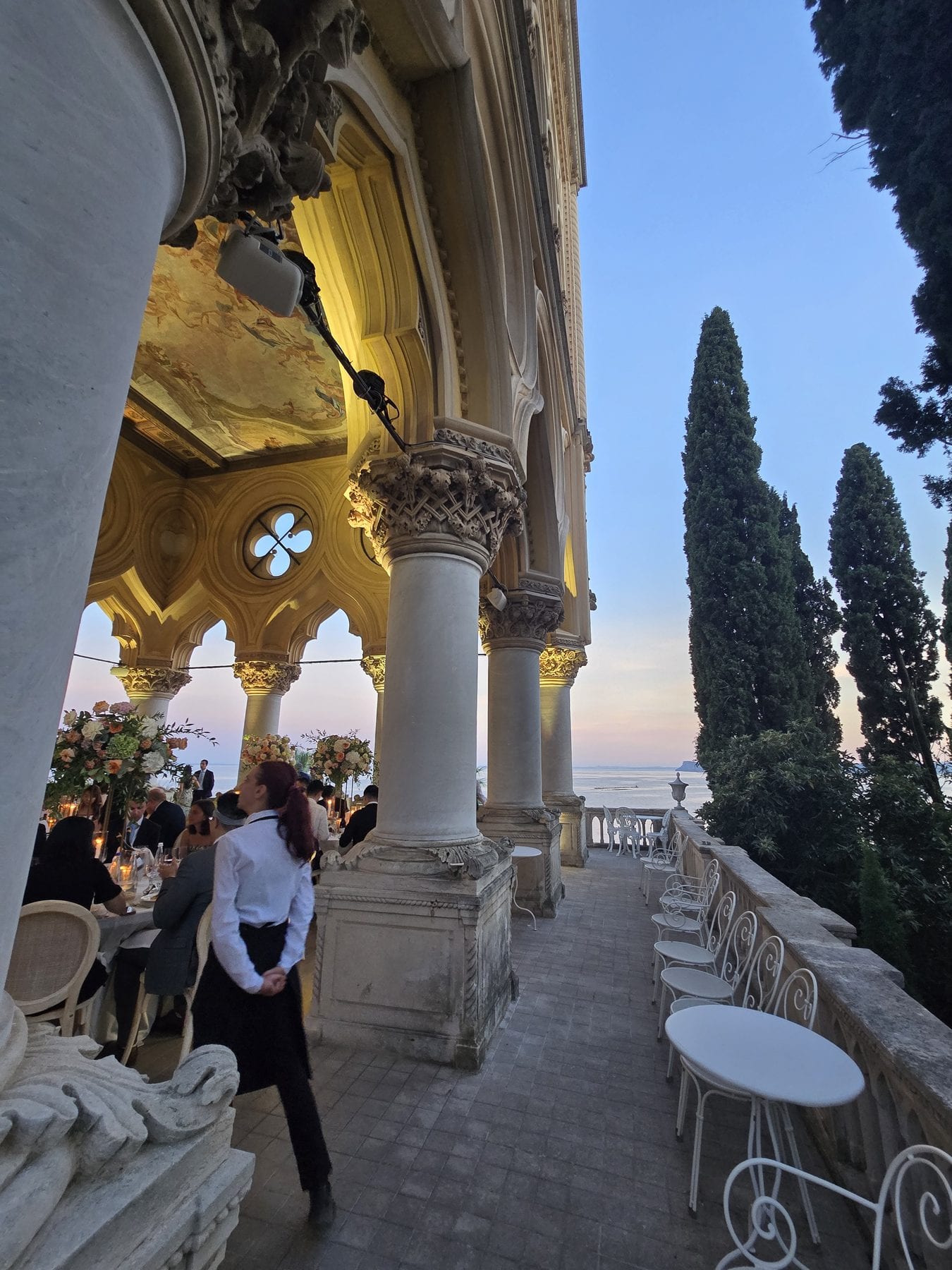 La loggia di una villa sull'Isola del Garda al tramonto: colonne, cipressi e i tavoli apparecchiati affacciati sul lago