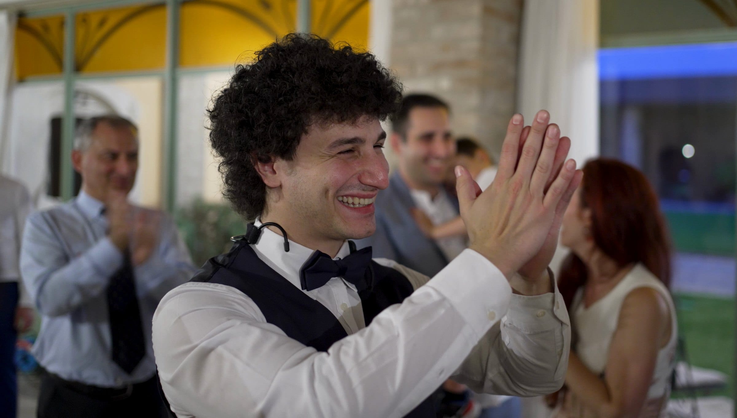 Matteo Angrisano sorride e applaude con gli ospiti durante un matrimonio, in camicia bianca, gilet e papillon