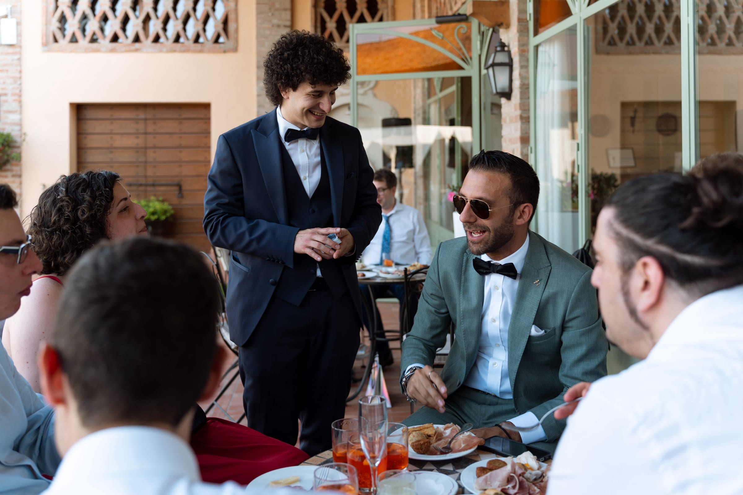Matteo Angrisano in smoking intrattiene gli invitati seduti al tavolo durante l'aperitivo di un matrimonio