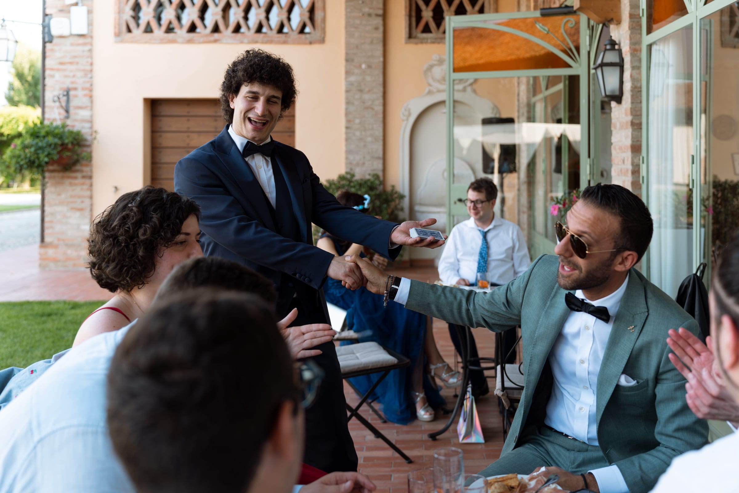 Matteo Angrisano stringe la mano a un ospite seduto al tavolo durante un ricevimento, mentre gli altri invitati applaudono