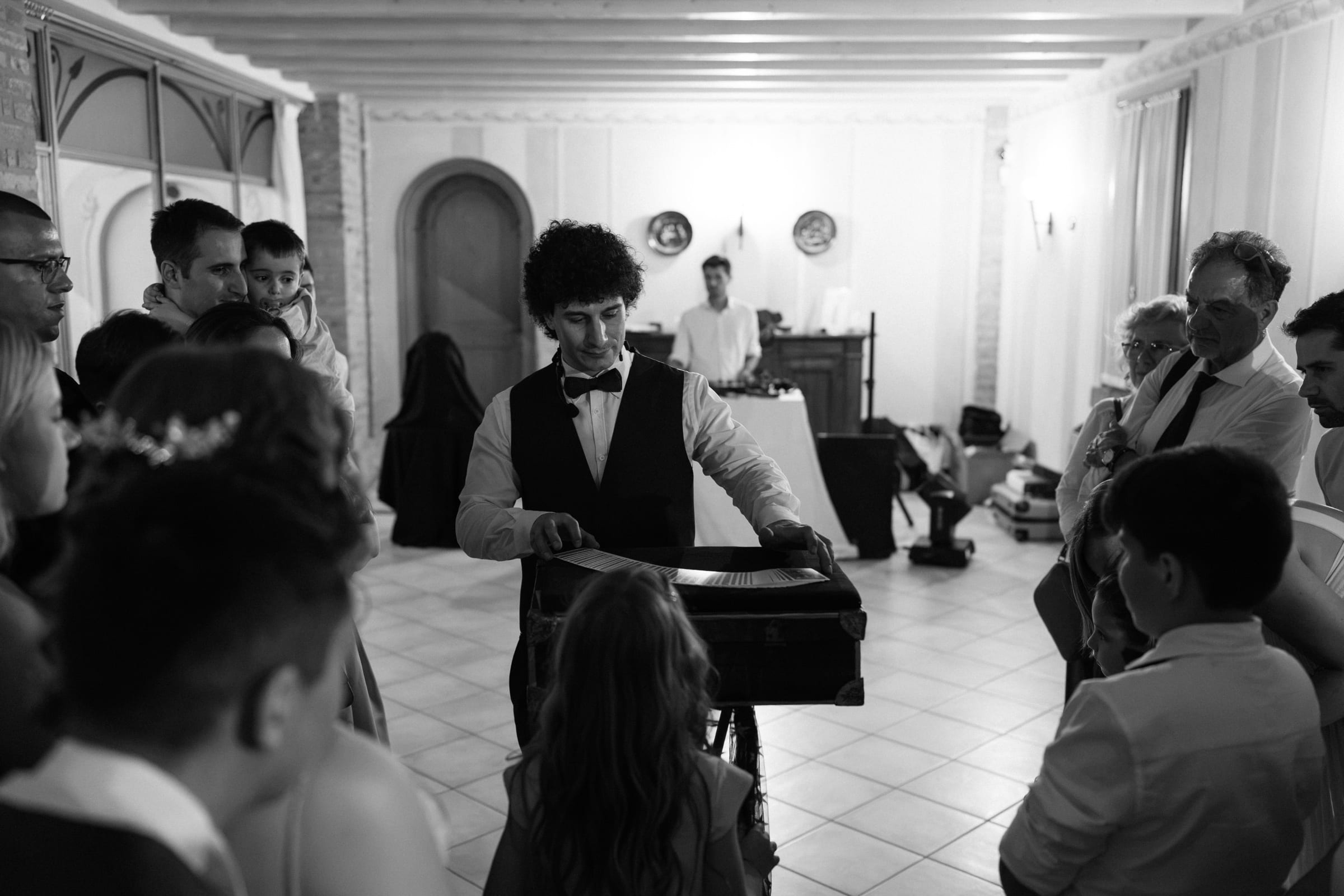 Fotografia in bianco e nero: gli ospiti di un matrimonio stretti attorno a Matteo Angrisano mentre esegue un gioco di carte