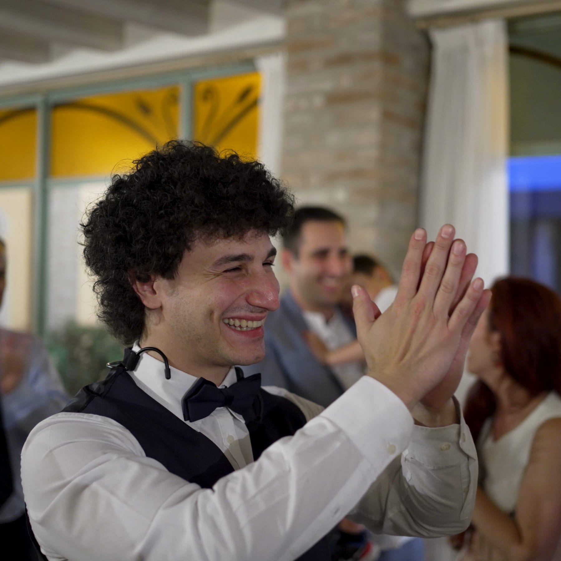Matteo Angrisano sorride e applaude con gli ospiti durante un matrimonio, in camicia bianca, gilet e papillon